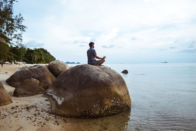 Rear view of man sitting on rock by sea against sky