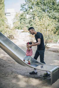 Young man holding hands of daughter walking on slide at playground