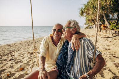 Happy senior gay man sitting with arm around boyfriend on rope swing at beach