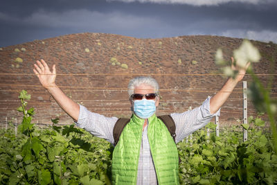 Portrait of man wearing mask while standing with arms raised amidst plants