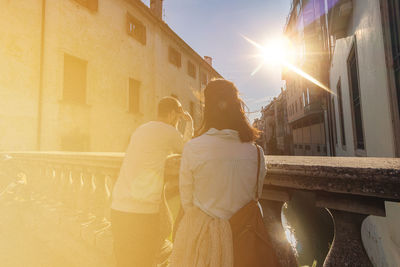 Rear view of couple standing by railing on footbridge over canal during sunny day