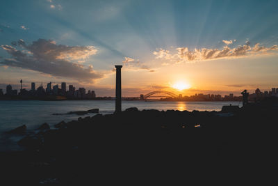 Silhouette buildings by sea against sky during sunset