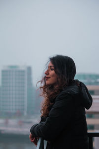 Side view of smiling young woman standing in city against sky 