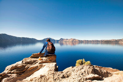 Man sitting on rock by lake against clear blue sky