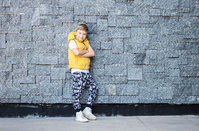 Full length portrait of boy with arms crossed standing against stone wall