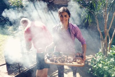 Woman carrying meat in tray while man standing by barbecue in yard