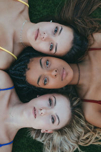 Top view of diverse female friends lying face to face on green lawn and looking at camera with serious expression