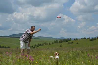 Man is playing with his dog on a meadow, surrounded with pink flowers and with hills in background