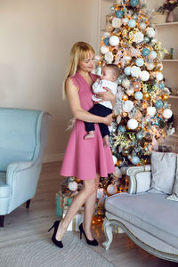 Woman in a pink dress holds her son's child in her arms standing at the christmas tree in the studio