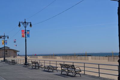 Empty park bench and street lights on pier at coney island against clear blue sky