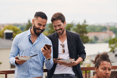 Smiling man showing mobile phone to friend while holding meal during party on terrace