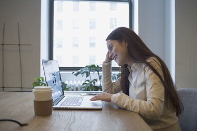 Young woman sitting at desk with coffee while looking at laptop computer.