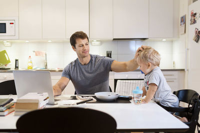 Father stroking baby boy while using laptop at dining table