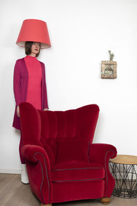 Mature woman standing behind armchair with lamp shade on head against white wall at home