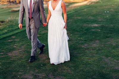 Low section of wedding couple holding hands while walking on field