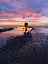 Camera on tripod at beach against cloudy sky during sunset