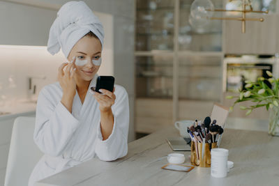 Smiling mother applying make-up while wearing bathrobe at home