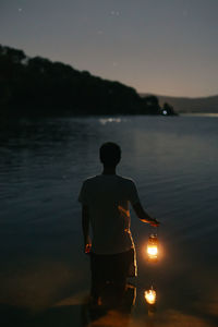 Back view of faceless male tourist with shiny lantern contemplating rippled river against mount at dusk