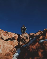 Low angle view of man sitting on rocky cliff against clear blue sky