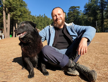 Full length of a eastern european man sitting outdoors with a black dog.
