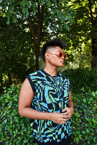 Filipino non binary person in stylish sunglasses with acne on face looking away while standing near tropical lush bush on summer day in park