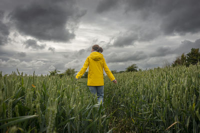 Woman in a yellow coat walking alone through a field, stormy, dramatic sky.