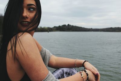 Close-up portrait of young woman sitting by lake against sky