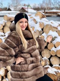 Portrait of smiling young woman wearing warm clothing by woodpile during winter