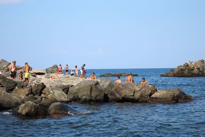 Group of people on rocks by sea against clear sky