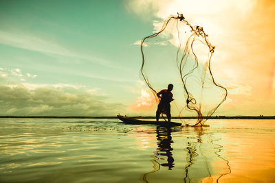 Man throwing fishing net in lake against sky during sunset