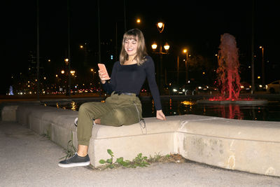Full length of young woman using smart phone while sitting outdoors at night
