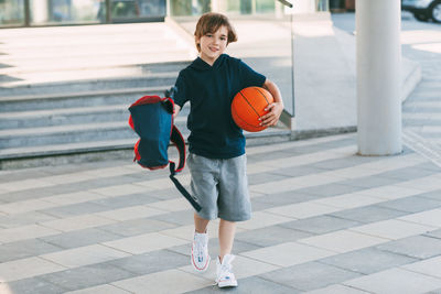 Portrait of boy holding basketball ball and backpack while walking on road