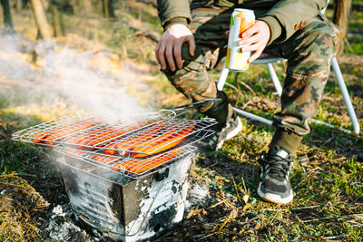 Person holding beer and watching barbecue grill in the forest
