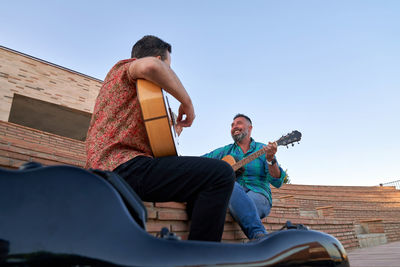 From below positive male musicians playing song on acoustic guitars while sitting together on steps on street of city