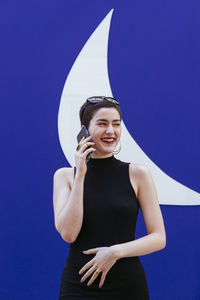 Smiling woman talking on phone while standing against blue background