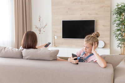 Girl looking at smart phone sitting by mother on sofa at home