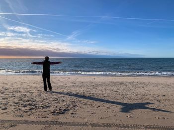 Rear view of woman with arms outstretched standing at beach against sky