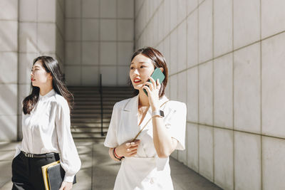 Coworker looking away while businesswoman talking on phone in city during sunny day