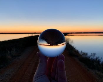 Cropped hand holding crystal ball by lake during sunset