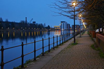 Footpath by river in city against sky at dusk
