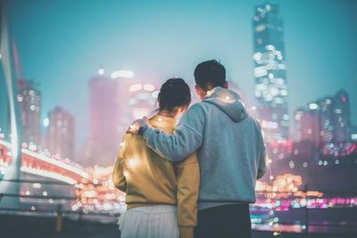 Rear view of couple looking at illuminated modern buildings while standing in city at night