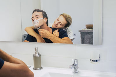 Man shaving beards while woman embracing at bathroom