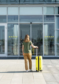 Young woman traveler carrying yellow suitcase next to the entrance to the airport outside tourism 