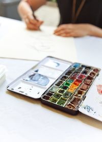 Midsection of woman sketching with colorful paints in foreground at table