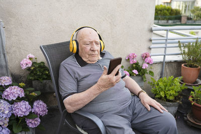 Senior man holding smart phone listening music through wireless headphones sitting on balcony