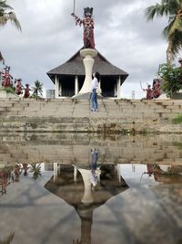 Reflection of woman on lake by building against sky