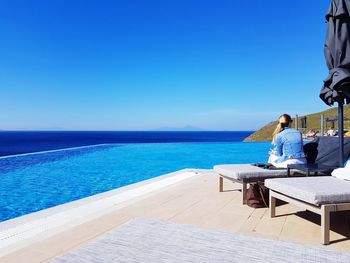 Rear view of woman sitting by infinity pool against clear blue sky during sunny day