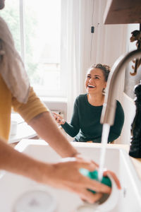 Cheerful woman looking at boyfriend washing utensils at home