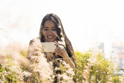 Smiling young woman photographing flowers with using mobile phone against clear sky