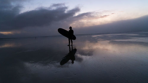 Silhouette young man with surfboard standing at beach against sky during sunset